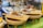 Bowls filled with delicious-looking stir-fried noodles, garnished with fresh lime and vegetables, are placed on woven bamboo stands. Wooden boards serve as trays supporting the bowls, suggesting an inviting and rustic presentation. The blurred background hints at a busy market or food stall atmosphere.