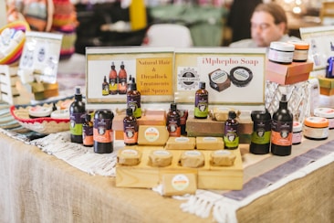 A market stall displaying an array of natural hair and skincare products, including bottles, jars, and soap bars. Promotional signs indicate offerings such as natural hair treatments, sprays, and soap. The products are neatly arranged on wooden blocks and layered fabric, with a variety of colors and branding labels visible.