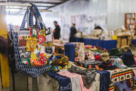 A vibrant and colorful tote bag featuring a patchwork of various designs, including a prominent lion's face, hangs on a display. The surrounding market scene includes multiple fabric items with diverse patterns and colors spread out on tables. In the background, blurred figures appear to be browsing through other stalls in a spacious indoor setting.