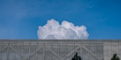 A large, billowing white cloud rises above a metallic mesh structure with some trees visible at the bottom edges. The sky is clear and blue, providing a stark contrast to the cloud and the structure.