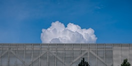 A large, billowing white cloud rises above a metallic mesh structure with some trees visible at the bottom edges. The sky is clear and blue, providing a stark contrast to the cloud and the structure.