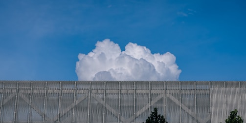A large, billowing white cloud rises above a metallic mesh structure with some trees visible at the bottom edges. The sky is clear and blue, providing a stark contrast to the cloud and the structure.