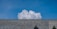 A large, billowing white cloud rises above a metallic mesh structure with some trees visible at the bottom edges. The sky is clear and blue, providing a stark contrast to the cloud and the structure.