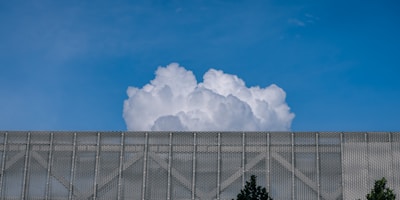A large, billowing white cloud rises above a metallic mesh structure with some trees visible at the bottom edges. The sky is clear and blue, providing a stark contrast to the cloud and the structure.