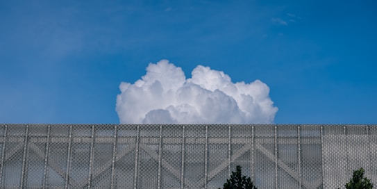 A large, billowing white cloud rises above a metallic mesh structure with some trees visible at the bottom edges. The sky is clear and blue, providing a stark contrast to the cloud and the structure.