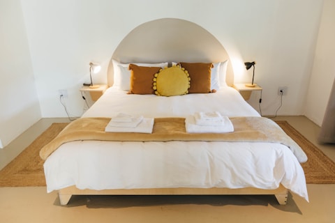 A neatly made bed in a minimalist bedroom setting. The bed has a white duvet, with a brown throw blanket and two white towels folded on it. There are three pillows, two in brown and one circular yellow pillow. Two bedside tables are on either side of the bed, each with a reading lamp. There is a semi-circular headboard against a beige wall and a rug underneath the bed.