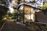 A modern glass-walled structure is nestled amidst lush greenery, with a stone path leading up to it. The structure features a pitched roof and appears to be a serene living space, flooded with natural light from the surrounding trees and garden. The area is tranquil and airy, blending contemporary architecture with nature.