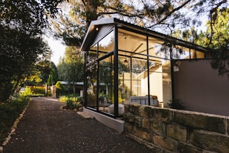 A modern glass-walled structure is nestled amidst lush greenery, with a stone path leading up to it. The structure features a pitched roof and appears to be a serene living space, flooded with natural light from the surrounding trees and garden. The area is tranquil and airy, blending contemporary architecture with nature.