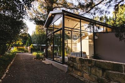 A modern glass-walled structure is nestled amidst lush greenery, with a stone path leading up to it. The structure features a pitched roof and appears to be a serene living space, flooded with natural light from the surrounding trees and garden. The area is tranquil and airy, blending contemporary architecture with nature.