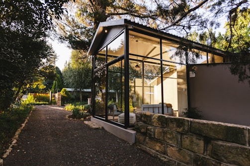 A modern glass-walled structure is nestled amidst lush greenery, with a stone path leading up to it. The structure features a pitched roof and appears to be a serene living space, flooded with natural light from the surrounding trees and garden. The area is tranquil and airy, blending contemporary architecture with nature.