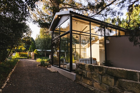 A modern glass-walled structure is nestled amidst lush greenery, with a stone path leading up to it. The structure features a pitched roof and appears to be a serene living space, flooded with natural light from the surrounding trees and garden. The area is tranquil and airy, blending contemporary architecture with nature.
