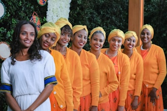 A group of eight women is standing in a line, smiling at the camera. They are dressed in vibrant outfits, with seven of them wearing orange cardigans, yellow headwraps, and patterned skirts. The woman on the left is in a white dress with blue and yellow accents. The background includes greenery and decorative, colorful elements on the wall.