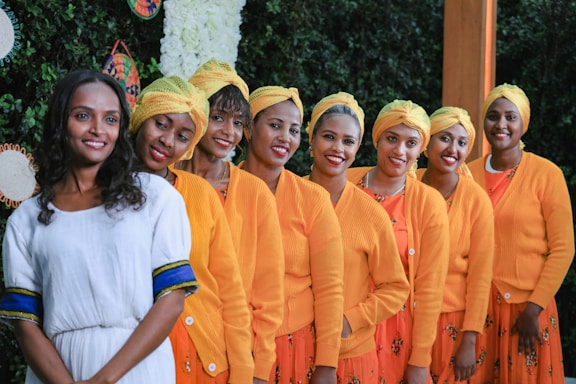 A group of eight women is standing in a line, smiling at the camera. They are dressed in vibrant outfits, with seven of them wearing orange cardigans, yellow headwraps, and patterned skirts. The woman on the left is in a white dress with blue and yellow accents. The background includes greenery and decorative, colorful elements on the wall.