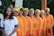 A group of eight women is standing in a line, smiling at the camera. They are dressed in vibrant outfits, with seven of them wearing orange cardigans, yellow headwraps, and patterned skirts. The woman on the left is in a white dress with blue and yellow accents. The background includes greenery and decorative, colorful elements on the wall.