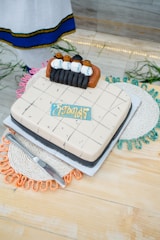A decorative cake features small figures of people in traditional attire placed on top, surrounded by detailed icing work. The cake rests on a light-colored wooden surface with colorful woven placemats below, and a metal knife is placed beside it.