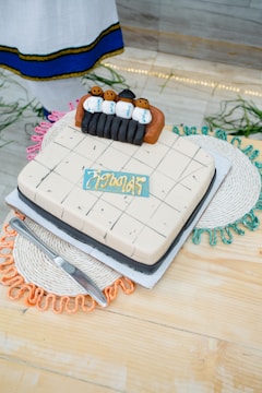 A decorative cake features small figures of people in traditional attire placed on top, surrounded by detailed icing work. The cake rests on a light-colored wooden surface with colorful woven placemats below, and a metal knife is placed beside it.
