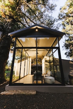 A glass-walled structure is situated among tall trees, featuring a modern design with visible furniture inside, such as sofas and cushions. The sunlight gently illuminates the space, creating shadows and reflections on the glass walls.