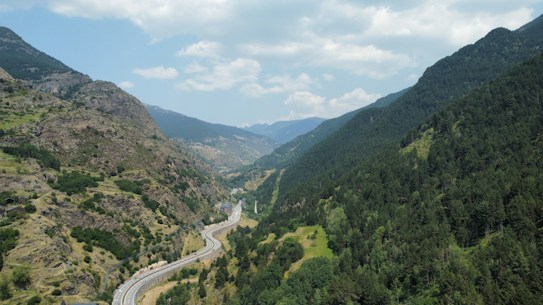 A winding road cuts through a lush green valley surrounded by towering mountains. The landscape features a mix of dense forests and rocky terrain, with a partly cloudy sky above adding a serene atmosphere.