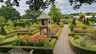 A beautifully landscaped garden featuring a variety of colorful flowers bordered by neatly trimmed hedges. In the center, there is a decorative structure resembling a small tower. The garden paths are gravel, and the area is surrounded by lush green trees. Benches and decorative elements are visible, enhancing the garden's aesthetic appeal.