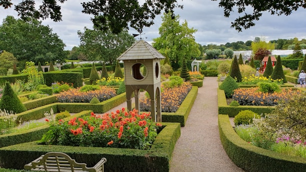 A beautifully landscaped garden featuring a variety of colorful flowers bordered by neatly trimmed hedges. In the center, there is a decorative structure resembling a small tower. The garden paths are gravel, and the area is surrounded by lush green trees. Benches and decorative elements are visible, enhancing the garden's aesthetic appeal.