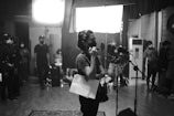 A black and white image depicts a busy sound studio. A person is standing near a microphone, seemingly speaking or preparing to speak. They hold papers in one hand and are wearing a mask. Several other individuals, also masked, can be seen in the background engaged in various activities. The room is equipped with professional lighting and sound equipment.