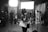 A black and white image depicts a busy sound studio. A person is standing near a microphone, seemingly speaking or preparing to speak. They hold papers in one hand and are wearing a mask. Several other individuals, also masked, can be seen in the background engaged in various activities. The room is equipped with professional lighting and sound equipment.