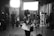 A black and white image depicts a busy sound studio. A person is standing near a microphone, seemingly speaking or preparing to speak. They hold papers in one hand and are wearing a mask. Several other individuals, also masked, can be seen in the background engaged in various activities. The room is equipped with professional lighting and sound equipment.