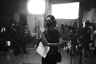 A group of people are gathered in a dimly lit recording studio. One person is standing in the foreground wearing headphones and holding papers, appearing focused. Several others are in the background, some wearing masks, busy with equipment and lights.