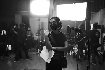 A group of people are gathered in a dimly lit recording studio. One person is standing in the foreground wearing headphones and holding papers, appearing focused. Several others are in the background, some wearing masks, busy with equipment and lights.