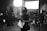 A group of people are gathered in a dimly lit recording studio. One person is standing in the foreground wearing headphones and holding papers, appearing focused. Several others are in the background, some wearing masks, busy with equipment and lights.