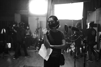 A group of people are gathered in a dimly lit recording studio. One person is standing in the foreground wearing headphones and holding papers, appearing focused. Several others are in the background, some wearing masks, busy with equipment and lights.