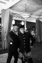 Two men dressed in formal wear are posing together in an elegant, dimly lit space with curtains and chandeliers. One man is wearing a suit, and the other is dressed in traditional attire with a hat. They appear to be in a jovial mood, with one gesturing towards his chin in a playful manner.