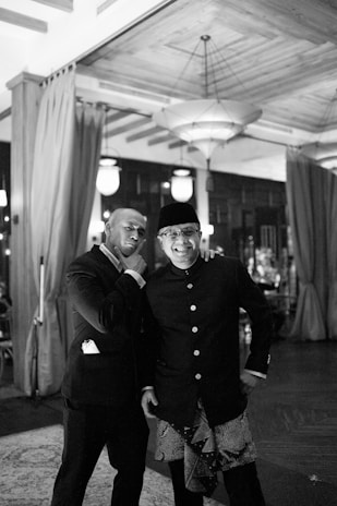 Two men dressed in formal wear are posing together in an elegant, dimly lit space with curtains and chandeliers. One man is wearing a suit, and the other is dressed in traditional attire with a hat. They appear to be in a jovial mood, with one gesturing towards his chin in a playful manner.