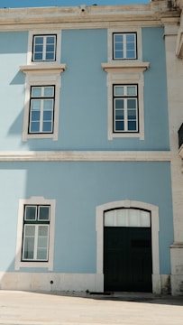 A facade of a building with a light blue wall and white trim. The building features two large windows on the upper story and one smaller window next to a large dark green door on the ground floor. The wall is illuminated by sunlight, casting slight shadows.