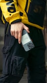 A person wearing a yellow jacket with bold black letters and black cargo pants is holding a smartphone. The jacket has a sporty, urban style, and the phone is held in a casual grip.