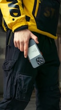 A person wearing a yellow jacket with bold black letters and black cargo pants is holding a smartphone. The jacket has a sporty, urban style, and the phone is held in a casual grip.