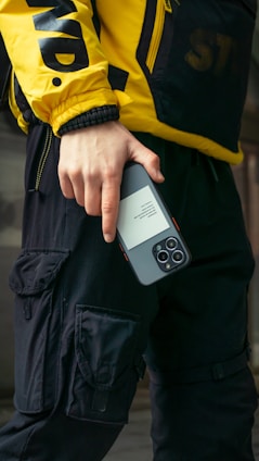 A person wearing a yellow jacket with bold black letters and black cargo pants is holding a smartphone. The jacket has a sporty, urban style, and the phone is held in a casual grip.