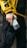 A person wearing a yellow jacket with bold black letters and black cargo pants is holding a smartphone. The jacket has a sporty, urban style, and the phone is held in a casual grip.
