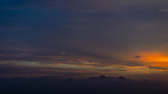 The image captures a serene sunset with dark mountains silhouetted beneath a sky filled with dramatic clouds. The horizon showcases a gradient of colors transitioning from dark blue to vibrant orange, with the sun casting warm hues across the scene.