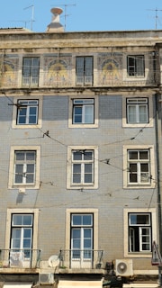 A multi-story apartment building features a facade adorned with intricate blue and white tiles, typical of Portuguese architecture. The upper level showcases decorative tilework with artistic designs, while the lower levels maintain a more uniform pattern. Several windows with white frames are symmetrically placed across the building. Air conditioning units are visible on the lower floors, and a variety of antennas extend from the rooftop. Electrical wires crisscross in front of the building, adding an urban texture to the scene.