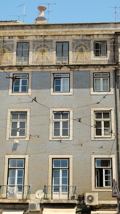 A multi-story apartment building features a facade adorned with intricate blue and white tiles, typical of Portuguese architecture. The upper level showcases decorative tilework with artistic designs, while the lower levels maintain a more uniform pattern. Several windows with white frames are symmetrically placed across the building. Air conditioning units are visible on the lower floors, and a variety of antennas extend from the rooftop. Electrical wires crisscross in front of the building, adding an urban texture to the scene.