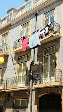 A multi-story building facade with decorative tiles and multiple balconies. Clothes are hanging out to dry on one of the balconies, revealing a variety of colors and fabrics. The windows have black wrought iron railings, and a bright, clear sky is visible above.