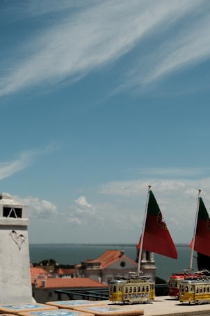 A scenic view with rooftops of orange tiles and historic buildings overlooking a large body of water under a sky with scattered clouds. In the foreground, there are small model trams adorned with Portuguese flags.