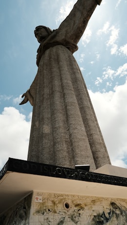 A large stone statue of a person with outstretched arms stands prominently against a partly cloudy sky. The base includes an inscription carved into the stone, reading 'God is Love'.