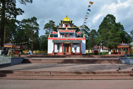 A colorful Buddhist temple with intricate architectural details stands at the center, surrounded by lush green trees. The building features multiple tiers with decorative elements in red, yellow, and blue. Prayer flags are strung above the temple, adding to the spiritual ambiance. The foreground consists of stone steps and paved ground leading up to the temple.