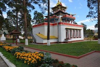 A traditional building with ornate architecture, featuring multiple levels with golden, red, and blue accents. Surrounding the structure is a well-maintained garden with vibrant orange flowers lining a pathway. Tall trees provide a peaceful, natural backdrop.