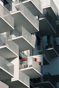 A modern apartment building with numerous balconies featuring metal railings. Some balconies display red flower pots with plants and shadows cast by the railing create geometric patterns across the building facade.