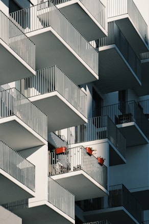 A modern apartment building with numerous balconies featuring metal railings. Some balconies display red flower pots with plants and shadows cast by the railing create geometric patterns across the building facade.