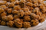 A large pile of cracked walnuts with their shells partially removed, revealing the wrinkled, cream-colored nuts inside. They are grouped on a white surface.