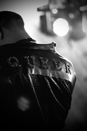 The image features a close-up view of a person from behind, wearing a jacket with the word 'QUEER' studded or embroidered on the back. The image is in black and white, with a focus on the texture of the jacket.